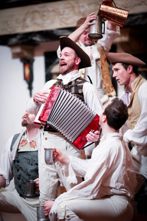 Sir Harry Bumper plays an accordian surrounded by the other four gallants in whit shirts and work vests and tri-cornered hats, all holding pewter mugs. 