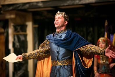 Pericles in tapestry shirt under dark blu waistcost with felt blue cape and gold crown, arms outstretched, the paper with the riddle in his right hand--Antiochus daughter is in the background. 