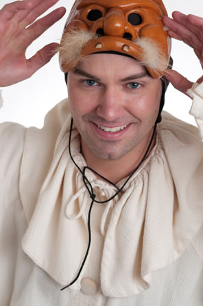 Wilson, smiling at the camera, has his hands reaching for the mask lying atop his head, a brown mask with pug nose, two fangs, and a tuft of white mustash poking out from above the lip.