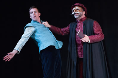 Shylock in a red quilt-like tunik,  black academic robe and red hat and wearing a brown mask with large white mustache and shaggy eyebrows holds a small account book in left hand and grabs the aqua blue tunic of Bassanio, who leans backward with arms spread.