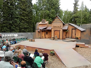 Photograph of the outdoor Fairbanks Shakespeare Theatre stage, with the wood facade Merry Wives set, ramps into the audience in chairs on the ground and platforms. Trees in the background