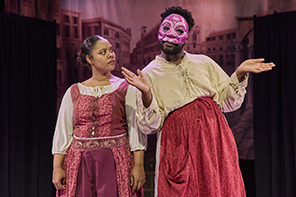 Juliet in red embroadered dress over white undershirt looks puzzled at Nurse, wearing a mask and red apron over white dress with her hands held out.