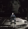 Hathaway in a flight suit sits on a chair in the middle of a gray-toned landscape with various coordinates on the edges, and a larger portrait of her projected on a screen in the background.