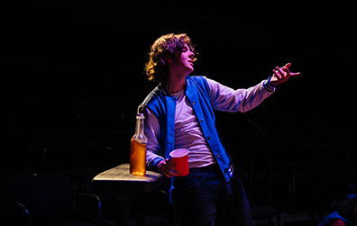 Duncan, wearing a blue and white letter jacket over a whit sweat shirt, gestureses with his left hand while holding a red cup in his right hand, his right elbow leaning on a counter on which is placed a long-neck bear. 