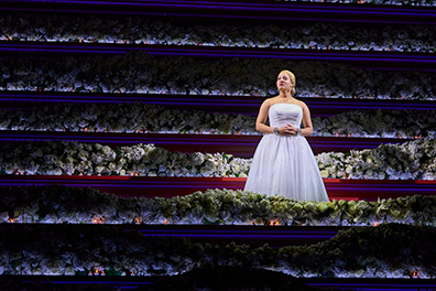 Under a spotlight, Eva Peron wearing a bared-shoulder, white ball gown, sings amid layers of candlit fwhite flowers.