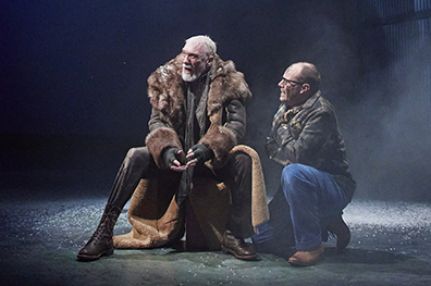 Patrick Page as King Lear sits on a case as Michael Milligan as the Fool kneels next to him. Lear is wearing a fur trimmed long coat, brown leather pants and boots and fingerless gloves. The Fool, folded arms against the cold, is wearing a camoflage jacket and blue jeans and glasses.Photo by DJ Corey Photography