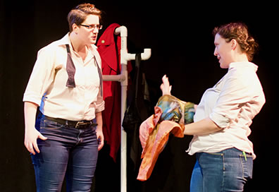 Production photo of Nicola Collett with an untied bowtie and Hannah Sweet a multicolored scarf The red coat hangs on piping in the background